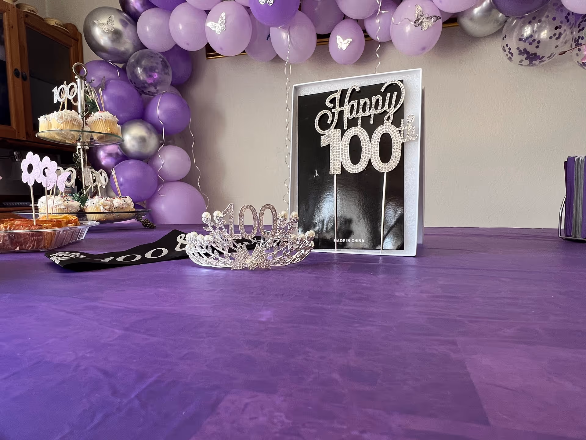 A decorated table with a purple tablecloth featuring a silver tiara with the number 100, a 'Happy 100th' sign, and a tiered tray with cupcakes and other treats. Purple and silver balloons with butterfly designs are arranged in an arch on the wall behind the table.