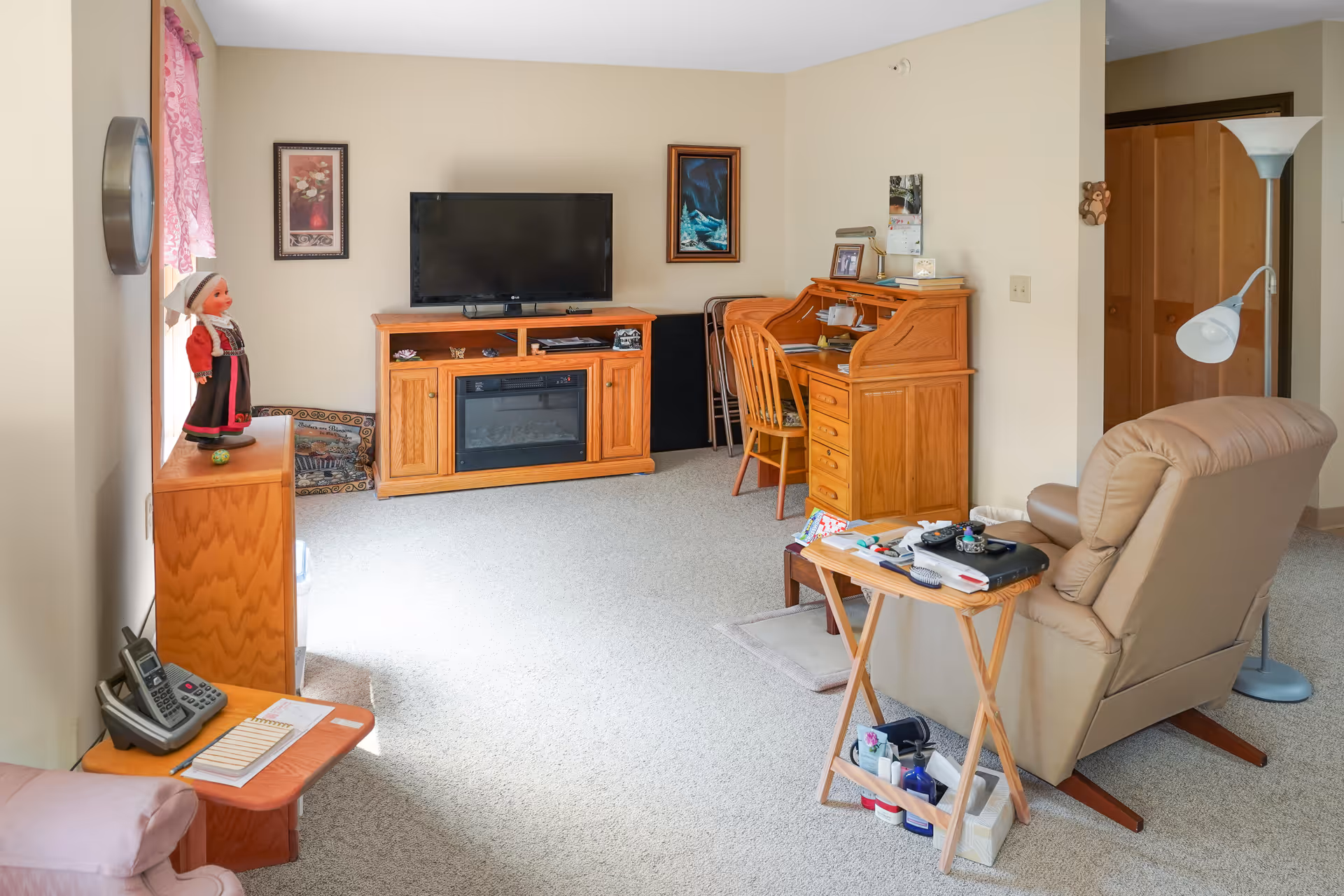 A cozy living room with beige walls and carpet. There is a wooden TV stand with a flat-screen TV and an electric fireplace. To the right, there is a wooden roll-top desk with a chair and some framed photos. A beige recliner chair faces a small wooden side table holding various items including a remote control and a lamp. On the left side, there is a small wooden cabinet with a doll dressed in traditional clothing on top. A window with pink curtains lets in natural light. A floor lamp stands near an open wooden door in the background.