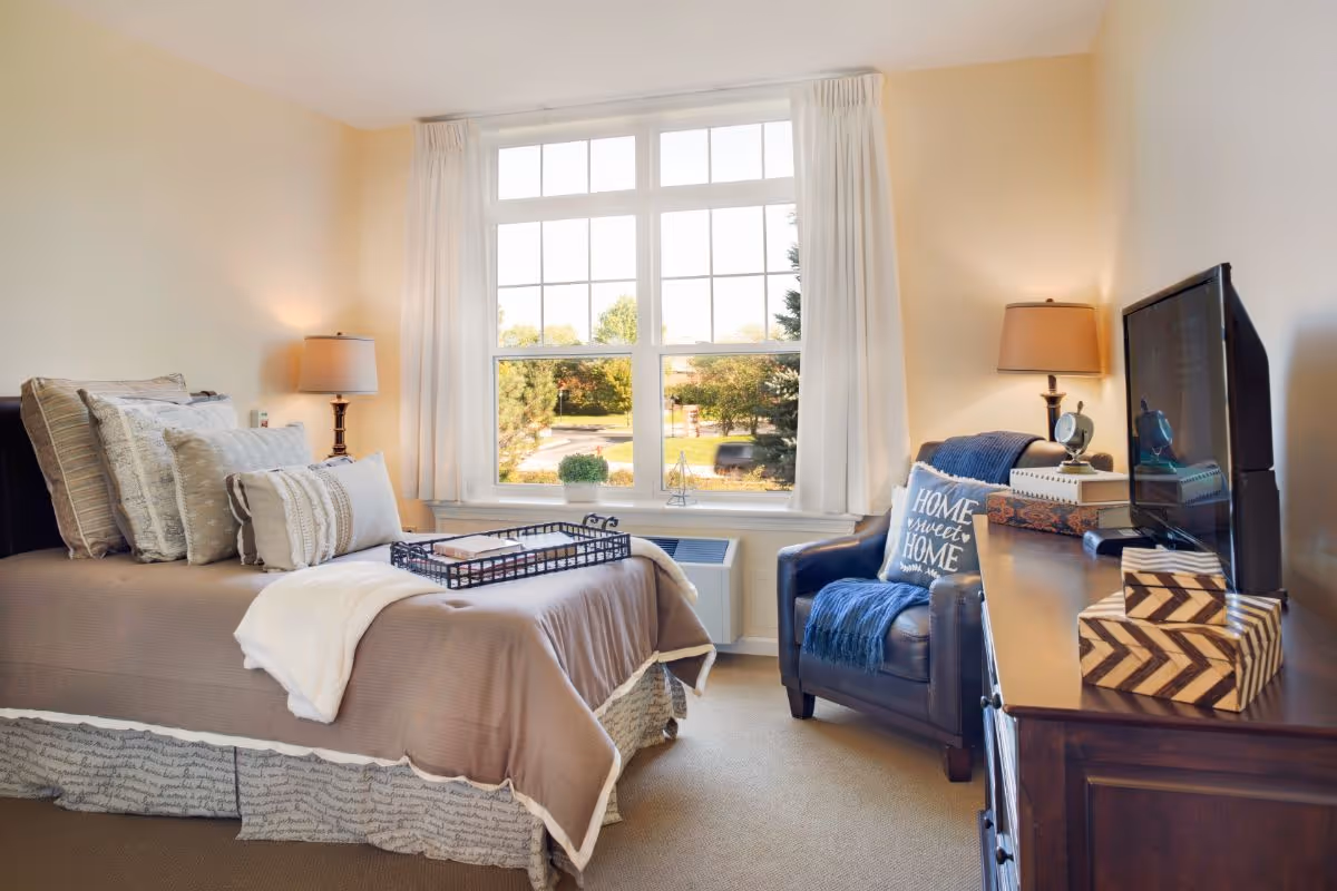 Bright, neatly furnished bedroom with a made bed, armchair, dresser and large window overlooking trees.