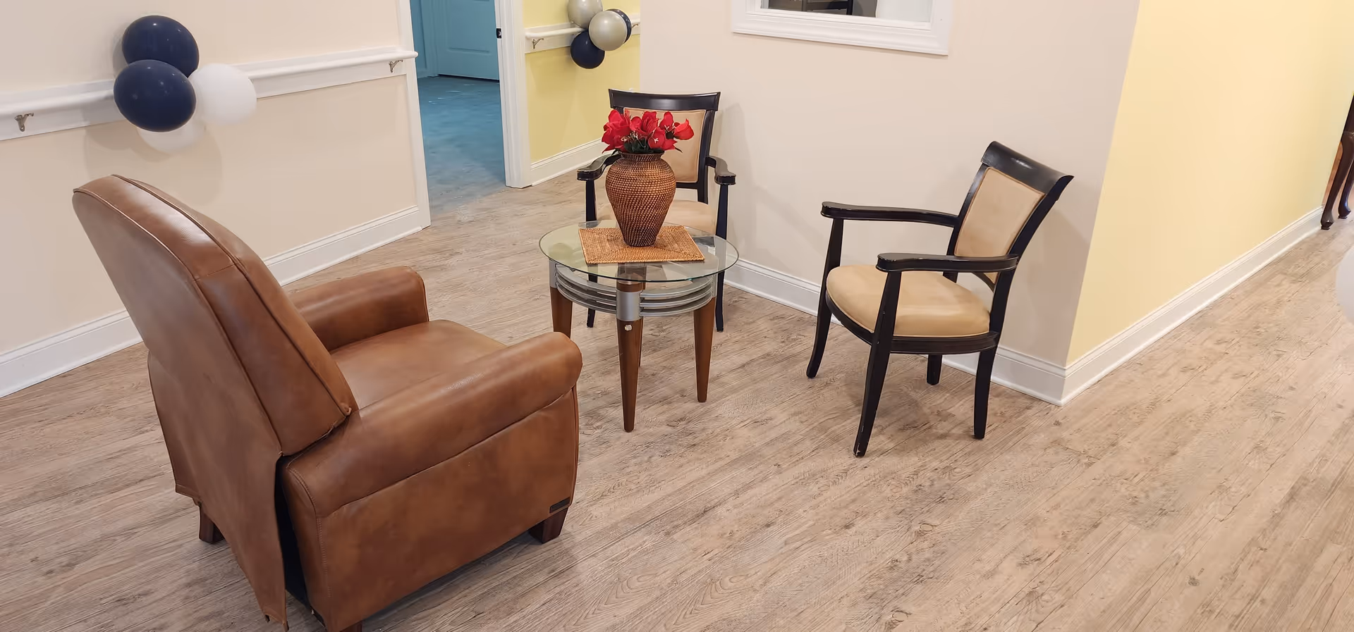 A cozy seating area in a senior care facility with a brown leather armchair, two wooden chairs with beige cushions, and a round glass coffee table with a vase of red flowers. The room has light-colored walls and wood flooring, with balloons attached to the wall in the background.