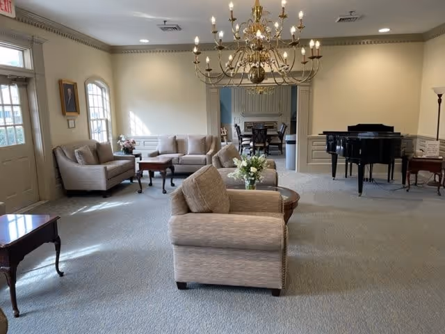 A spacious living room area with beige upholstered sofas and armchairs arranged around wooden coffee tables. A large chandelier hangs from the ceiling. In the background, there is a black grand piano and a dining area with a table and chairs. The room has light-colored walls and carpeted flooring, with natural light coming through windows on the left side.