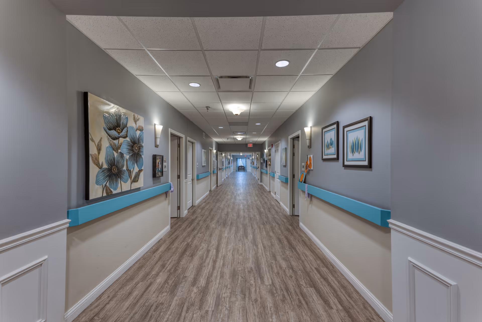 A long, well-lit hallway in a senior living facility with light gray walls, wood-patterned flooring, and blue handrails on both sides. The hallway is decorated with framed artwork and wall sconces, and multiple doors line each side.