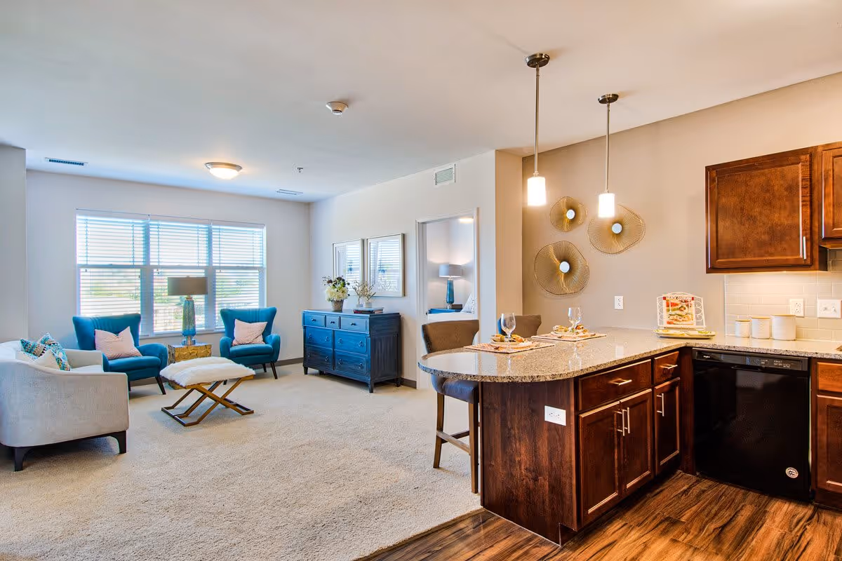 A bright and spacious living area in a senior living facility featuring a sitting area with two blue armchairs, a beige sofa, a small ottoman, and a blue sideboard under two framed pictures. Adjacent to the living area is a kitchen with dark wood cabinets, a granite countertop island with two place settings, and modern pendant lights hanging above. A bedroom is visible through an open doorway.