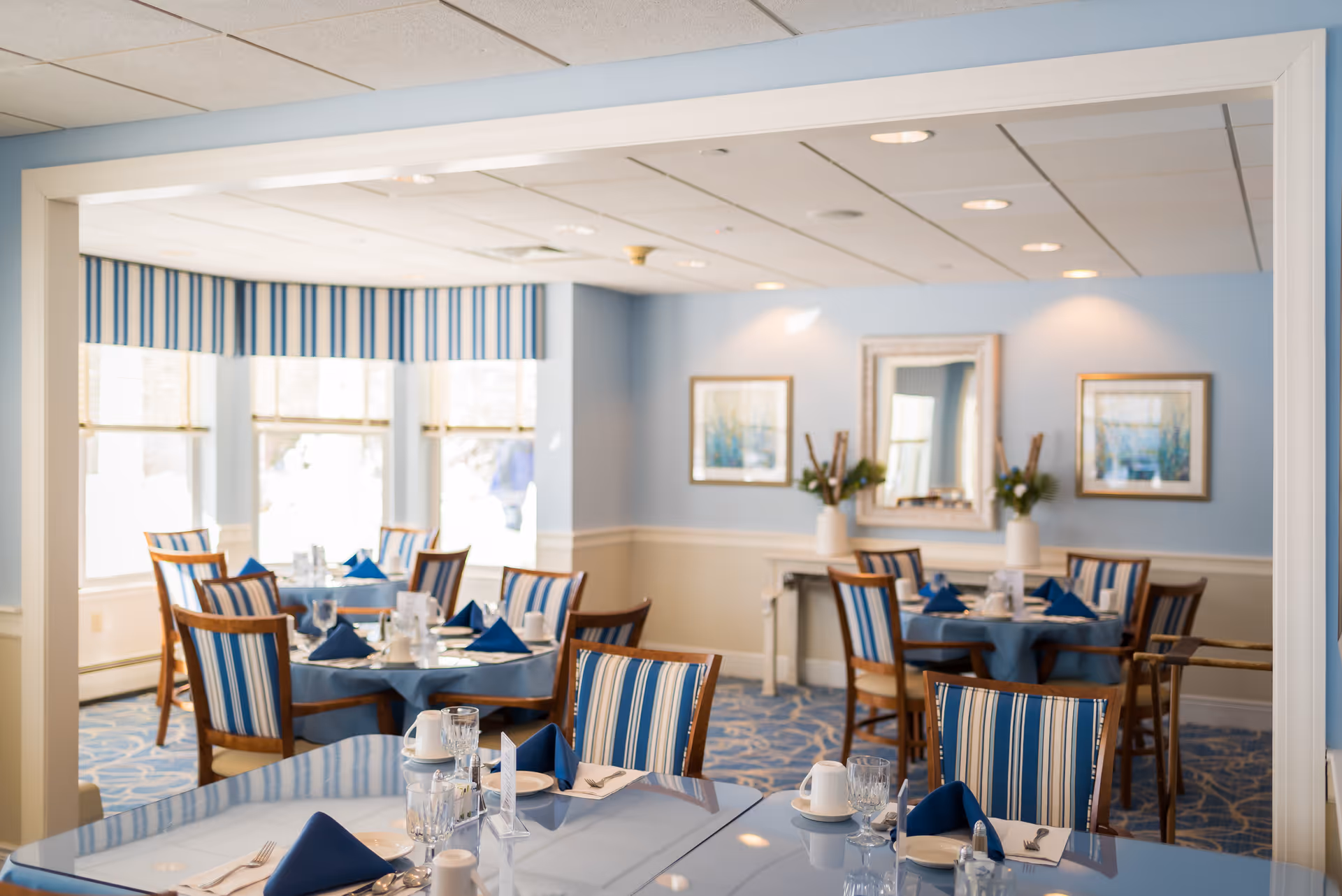 Well-lit dining room with round tables set with blue napkins and striped chairs.