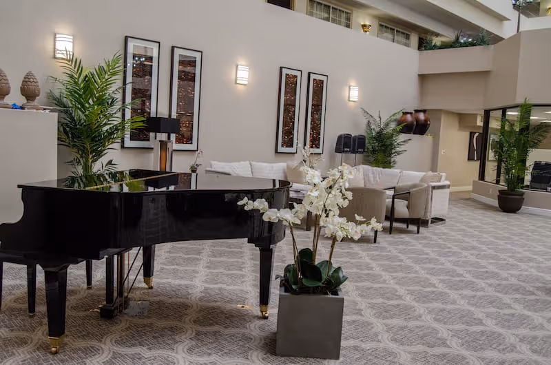 Spacious and elegant senior living community common area featuring a black grand piano, white cushioned sofas, armchairs, decorative plants, and framed wall art under soft wall lighting.