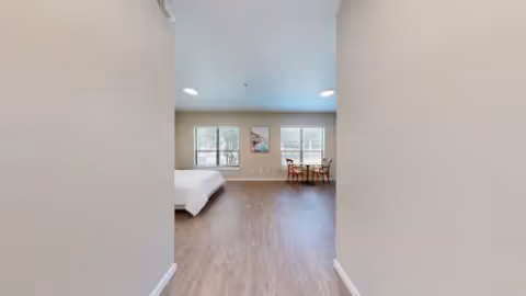 View through a hallway into a spacious bedroom with two large windows, a bed on the left side, and a small table with two chairs near the windows. The room has wood flooring and neutral-colored walls.