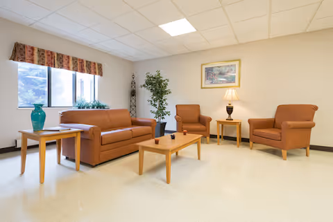 Bright communal sitting area with a sofa, two armchairs, coffee and side tables, a lamp and potted plants near a window.
