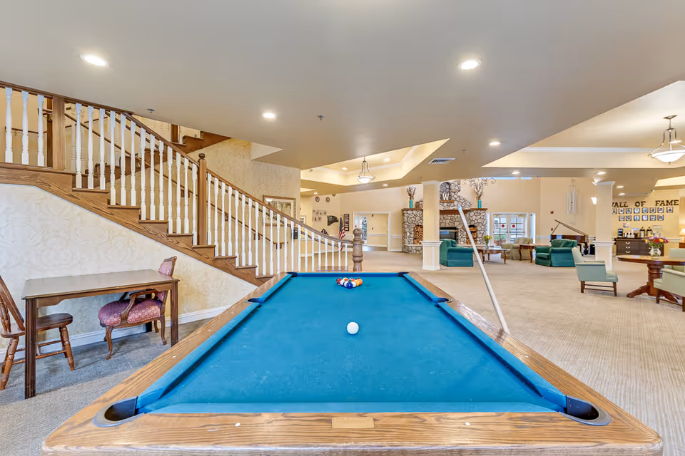 Interior view of a senior living facility common area featuring a pool table with a cue stick resting on it, a wooden staircase with white balusters on the left, several seating areas with green and beige chairs, a stone fireplace in the background, and a 'Wall of Fame' display on the far wall.