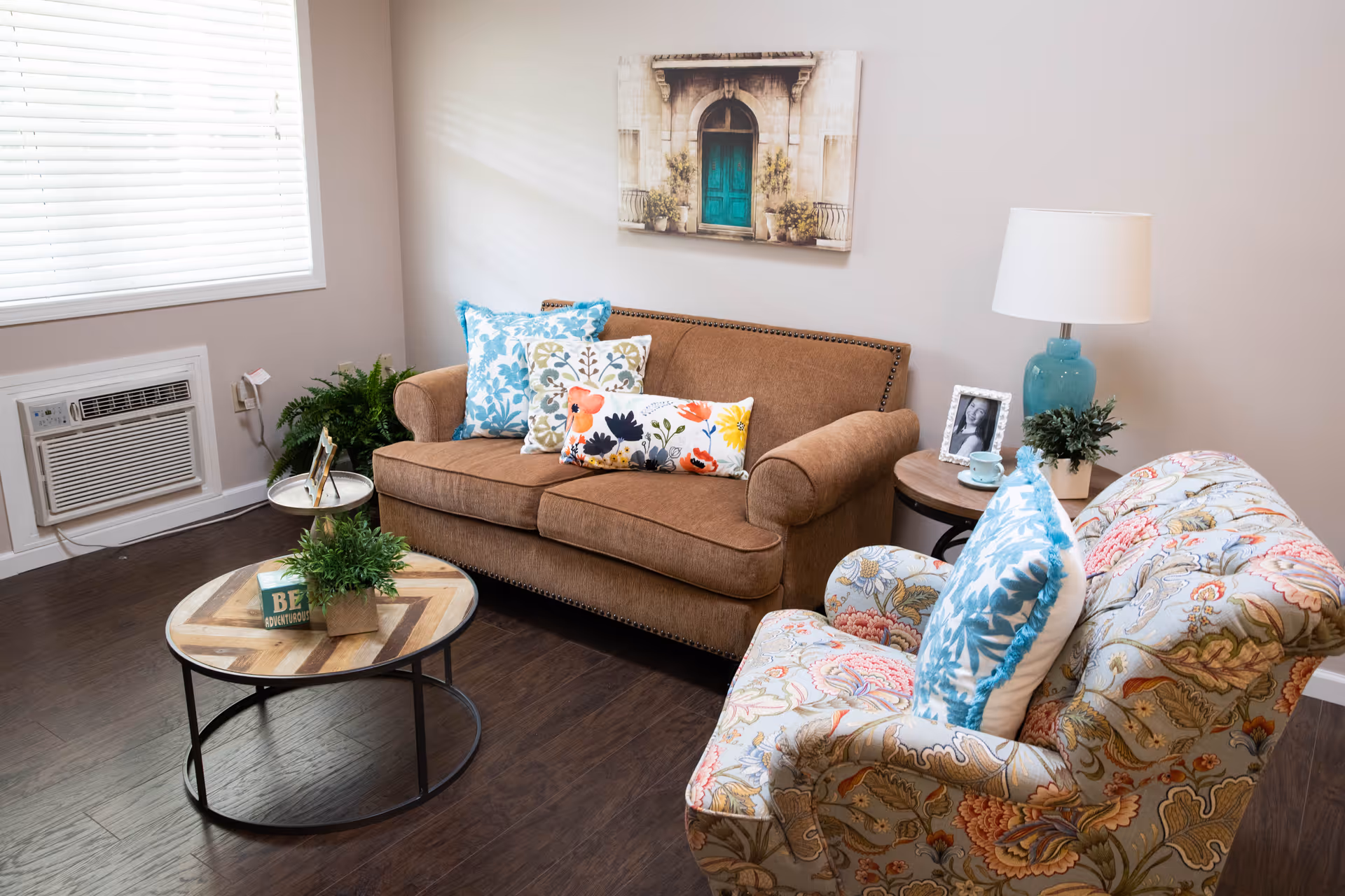 A cozy living room with a brown sofa adorned with colorful floral and patterned pillows, a patterned armchair with a blue and white pillow, a round wooden coffee table with a small plant and a decorative block, a side table with a blue lamp, a framed photo, and a small plant. There is a window with white blinds and an air conditioning unit below it. A painting of a door with plants hangs on the wall above the sofa.