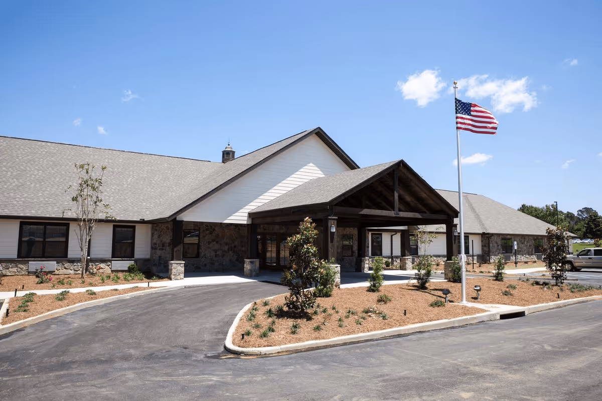 Exterior view of The Aspen of Brookhaven facility showing a single-story building with a stone and white siding facade, a covered entrance supported by wooden beams, a landscaped area with small plants and trees, an American flag on a flagpole, and a clear blue sky.