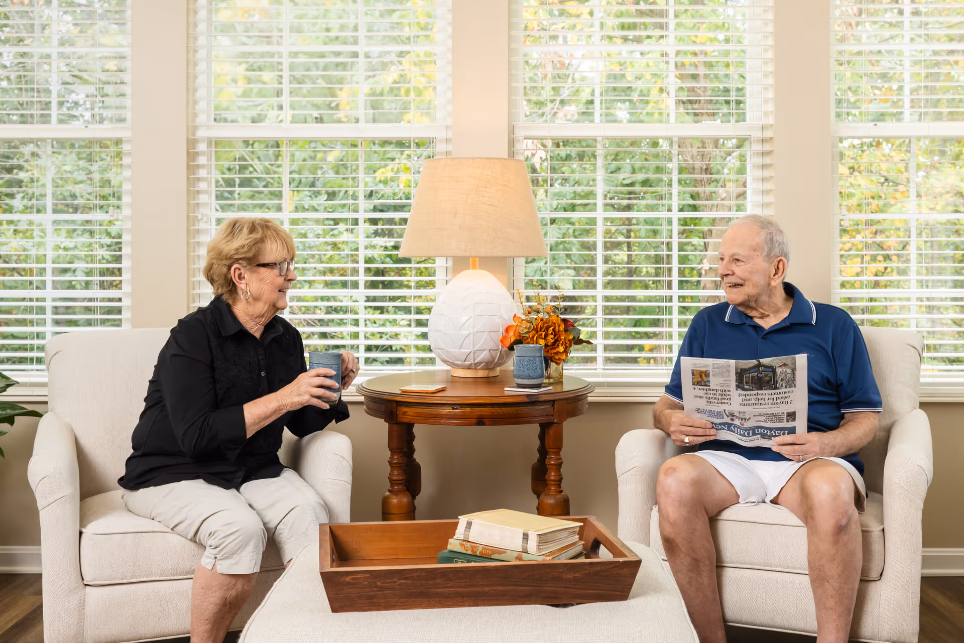 An elderly woman and an elderly man sitting in armchairs facing each other in a bright room with large windows and white blinds. The woman is holding a cup and smiling, while the man is holding a newspaper and smiling back. Between them is a wooden side table with a lamp and a small flower arrangement. In front of them is a wooden tray with books on a cushioned ottoman.