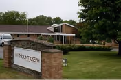 Exterior view of Mount Joseph Senior Village showing a brick sign with the facility name in front of a single-story building surrounded by green grass and trees under a cloudy sky.