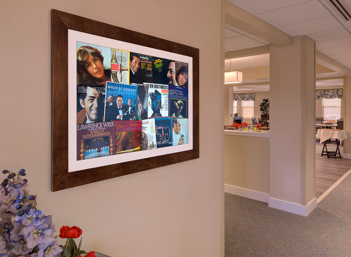 A framed collage of vintage album covers hangs on a hallway wall with a view into a dining area of the facility.