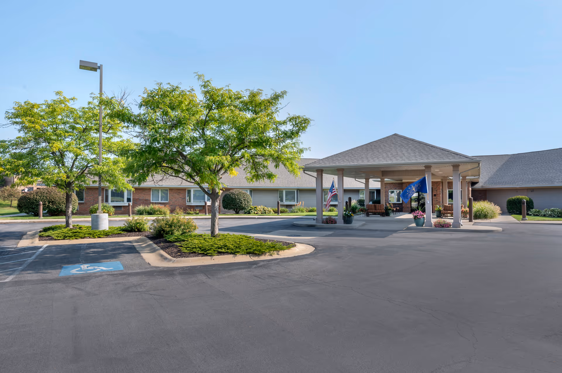Exterior view of a single-story senior living facility with a covered entrance, two flags, and landscaped greenery including trees and bushes. The parking lot in front includes a handicapped parking space.