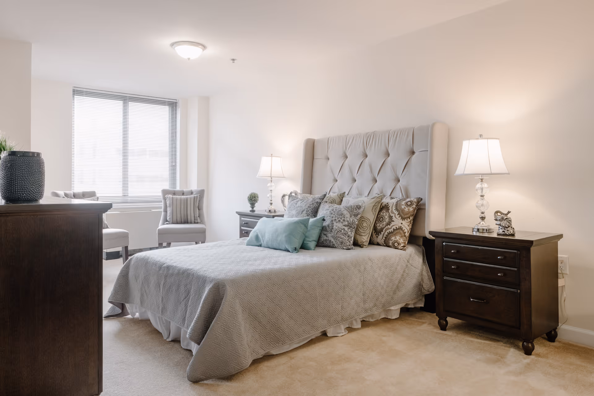 A well-lit bedroom with a large upholstered bed adorned with multiple decorative pillows. There are two dark wooden nightstands on either side of the bed, each with a lamp. Two cushioned chairs are positioned near a window with blinds, and a dark wooden dresser is visible in the foreground.