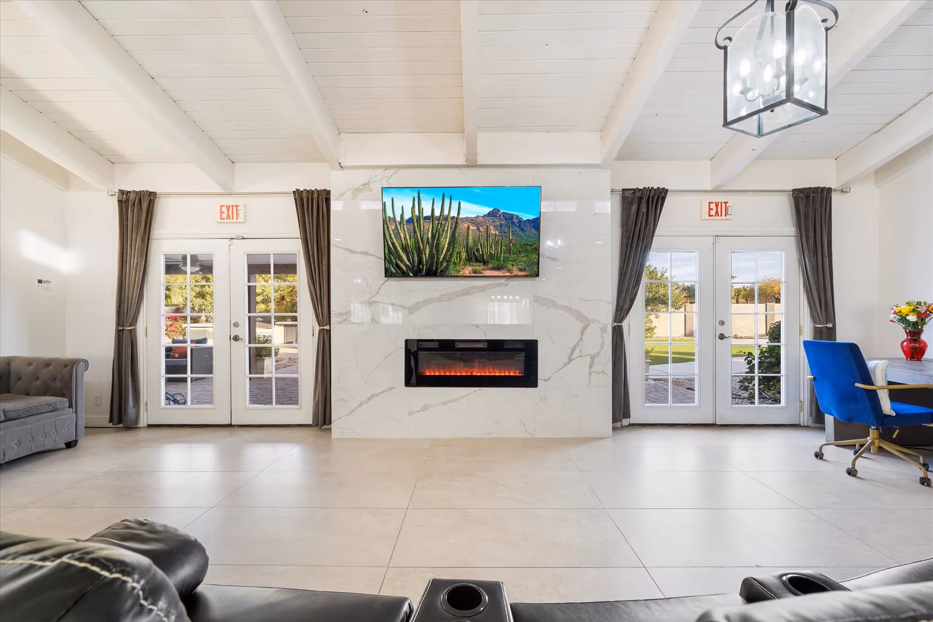 A bright and spacious living room with white walls and ceiling beams, featuring a wall-mounted flat-screen TV above a modern electric fireplace set in a marble surround. There are two sets of French doors with glass panes and dark curtains on either side of the fireplace, leading to an outdoor area. The room includes a gray tufted sofa on the left and a blue office chair with a desk and a red vase with flowers on the right. The floor is tiled in a light color, and there are exit signs above each set of doors.