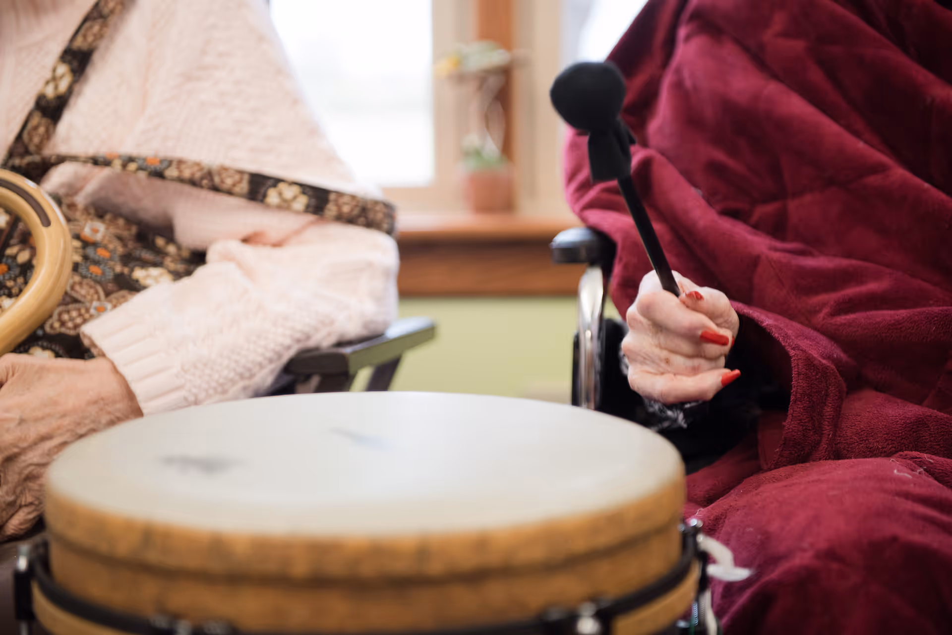 Close-up of two elderly individuals sitting side by side, one holding a drumstick and playing a drum, both wearing cozy clothing with a window and potted plant in the background.
