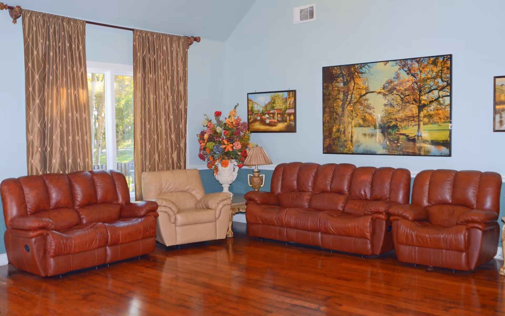 A living room with three brown leather sofas and one beige armchair arranged along light blue walls. There are three framed paintings on the wall, a large floral arrangement in a white vase on a side table, and a window with brown patterned curtains letting in natural light. The floor is polished wood.