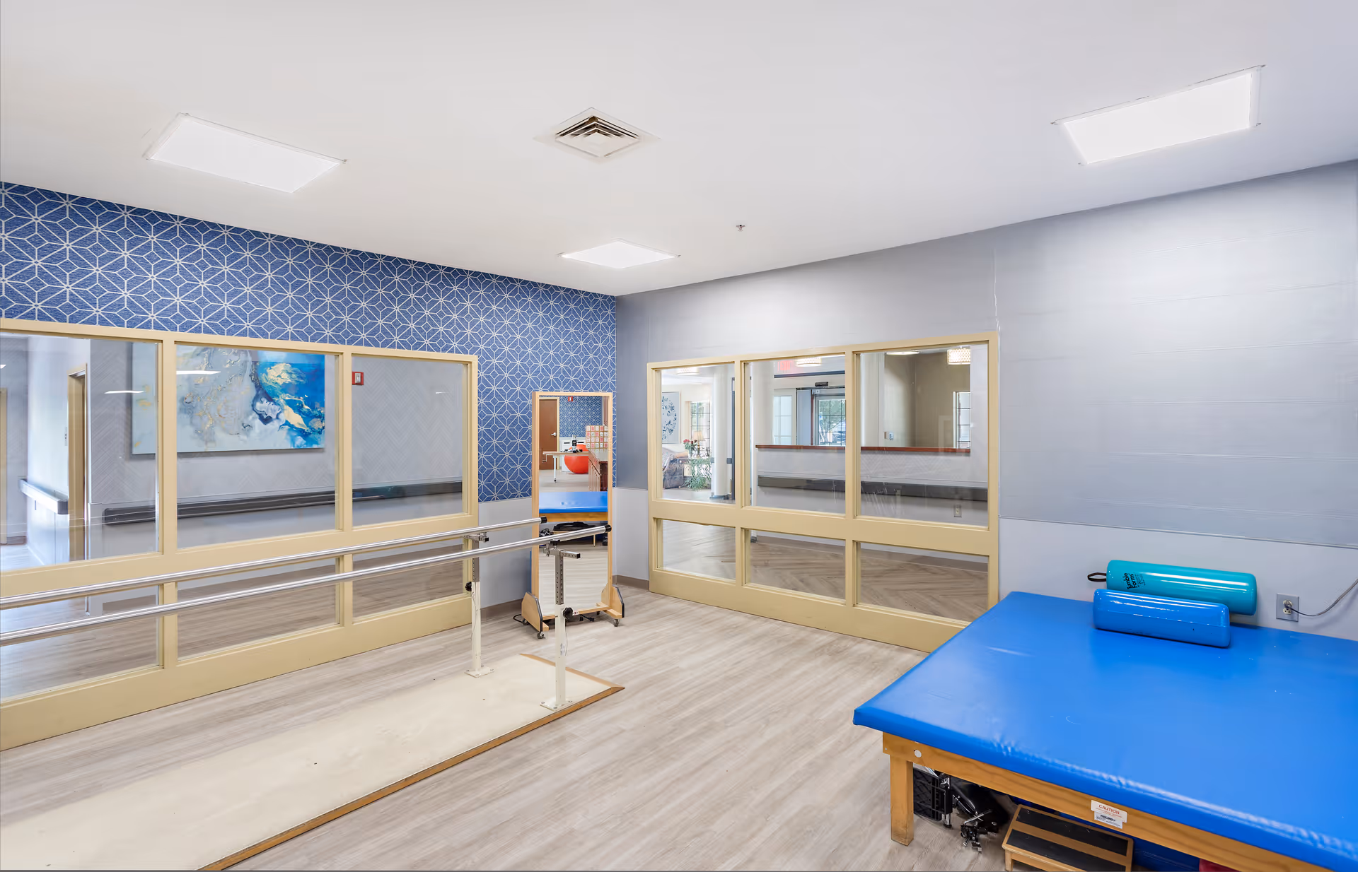Bright indoor rehabilitation room with parallel walking bars, a blue padded therapy table, and large interior windows.