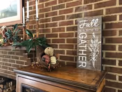 A rustic wooden cabinet against a brick wall with decorative items on top including a basket with pine cones and ornamental balls, a plant, two tall white candles in holders, and a wooden sign that reads 'Fall is a time to GATHER' with a wheat design.