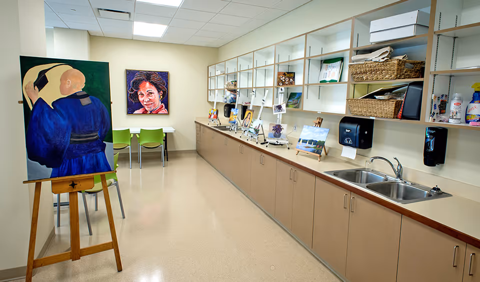 A bright art studio room with a long counter and cabinets along one wall, featuring a double sink, shelves with baskets and art supplies, and several small paintings displayed on easels. There is a large painting on an easel in the foreground and a portrait painting hanging on the far wall above two green chairs and a small table.