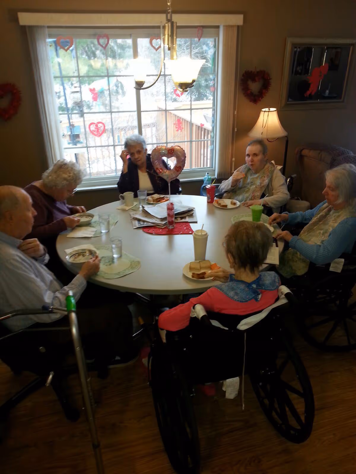 Several elderly residents seated around a round dining table in a decorated common room, eating and talking.