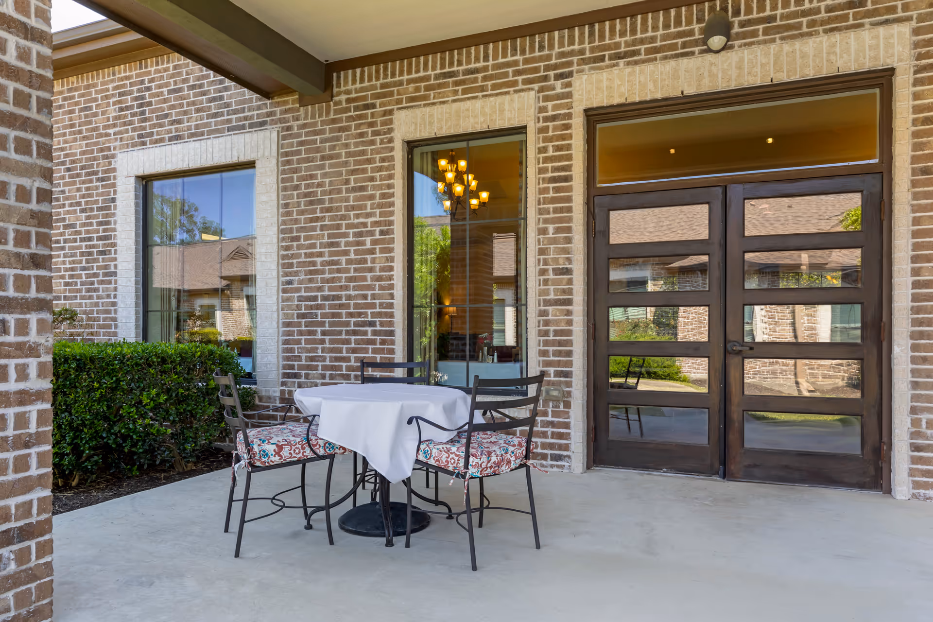 Outdoor patio area at Ansel Park Assisted Living & Memory Care featuring a round table covered with a white tablecloth and four metal chairs with patterned cushions. The patio is adjacent to a brick building with large windows and a double wooden door with glass panels.