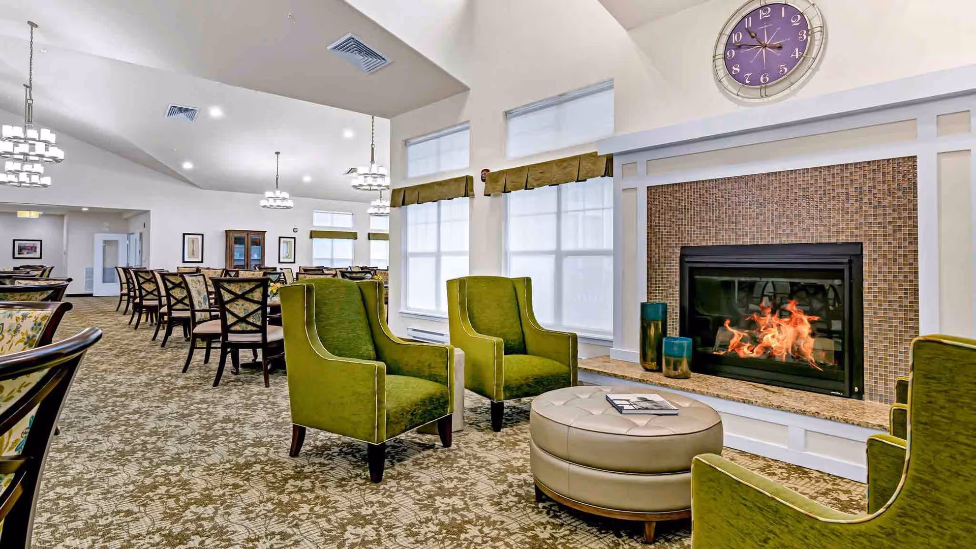 A spacious and well-lit common area with a fireplace surrounded by mosaic tiles. In front of the fireplace are four green upholstered armchairs arranged around a round beige ottoman with a book on top. The room has large windows with green valances, patterned carpet, and multiple dining tables with chairs in the background. Modern chandeliers hang from the vaulted ceiling.