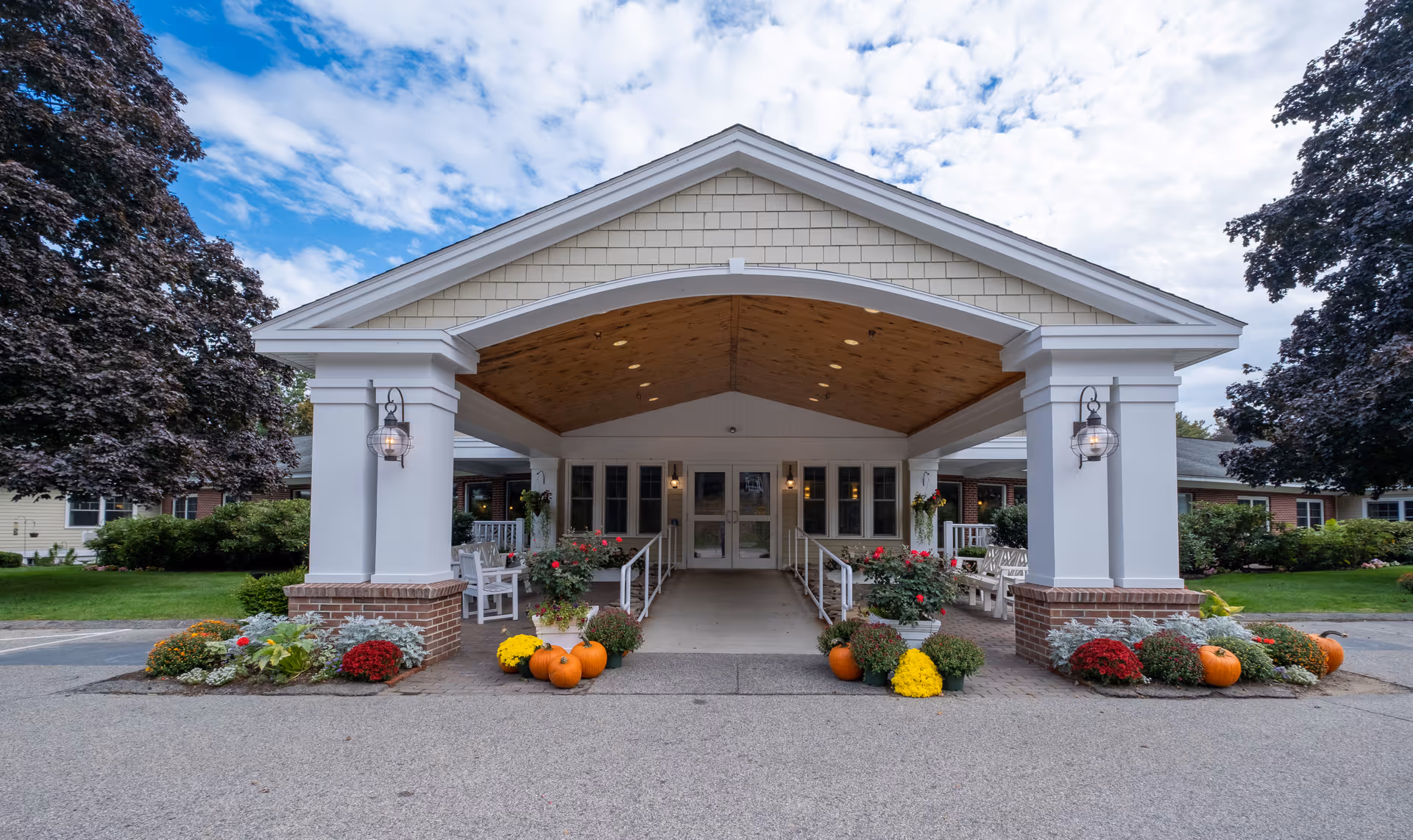 Front entrance of a senior living facility with a covered driveway supported by white columns. The entrance is decorated with pumpkins and colorful flowers on both sides. There are benches and greenery around the entrance, with a partly cloudy sky above.
