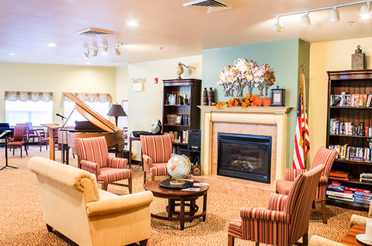 A cozy living room area in Rivercrest Place featuring a beige couch, four striped armchairs arranged around a round wooden coffee table with a globe on it, a fireplace decorated with autumn-themed ornaments, two bookshelves filled with books and games, an American flag, and a grand piano in the background near windows with valances.