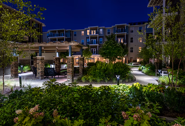 Night view of a senior living facility courtyard with well-lit walking paths, lush green landscaping, trees, and a covered seating area with chairs and tables. The multi-story building with balconies is visible in the background under a dark blue sky.