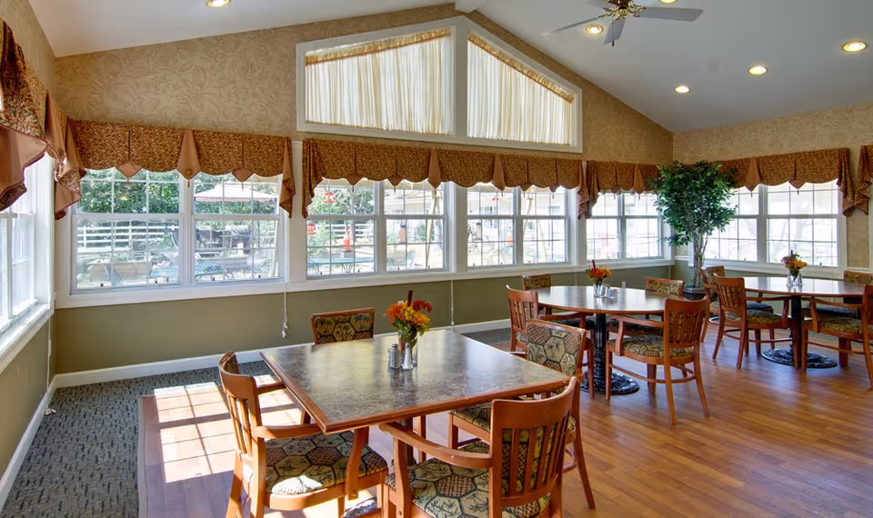 A bright dining room with several wooden tables and chairs, each table decorated with a small vase of flowers. Large windows with patterned valances let in natural light, and a tall potted plant is placed near the corner. The room has wood flooring and a high ceiling with recessed lighting and a ceiling fan.