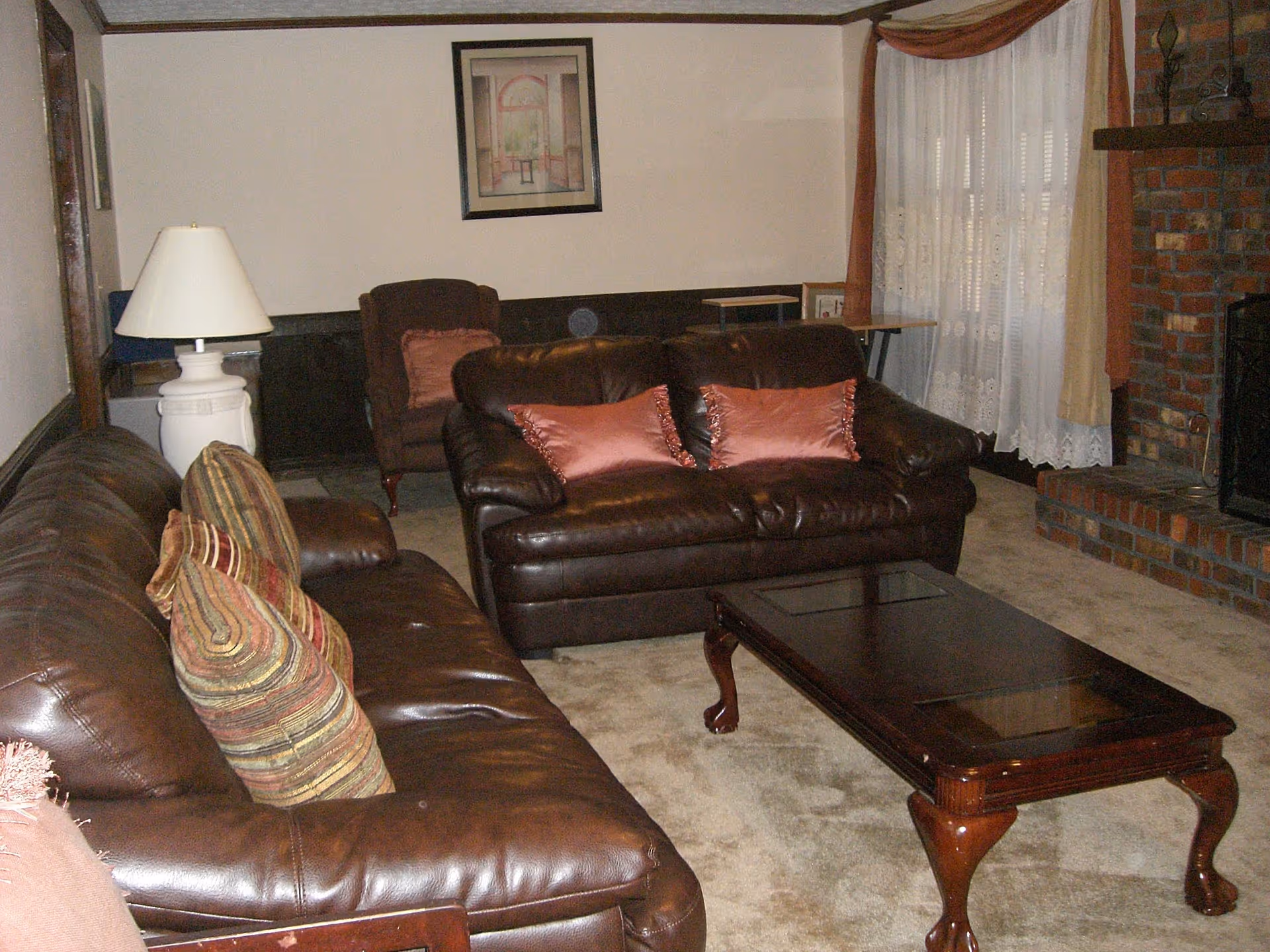 A cozy living room with two brown leather sofas adorned with decorative pillows, a wooden coffee table with glass inserts, a white table lamp on a side table, a framed picture on the wall, and a brick fireplace with a window covered by lace curtains and drapes.