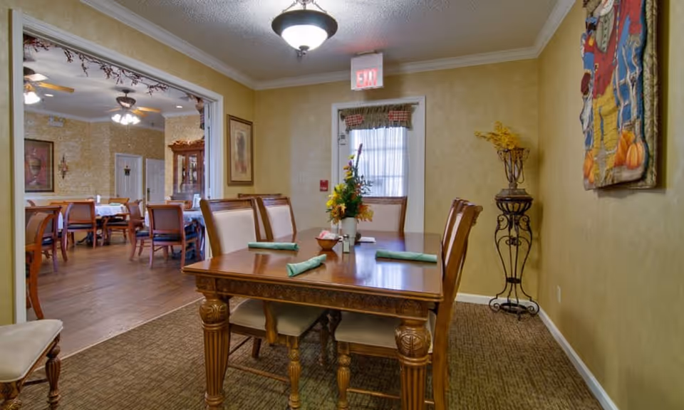 A dining area in a senior living facility with a wooden table set for four, each place setting with a green napkin. The room has beige walls, a carpeted floor, and a decorative flower arrangement on the table. In the background, there is an adjoining larger dining room with multiple tables and chairs, ceiling fans, and framed artwork on the walls.