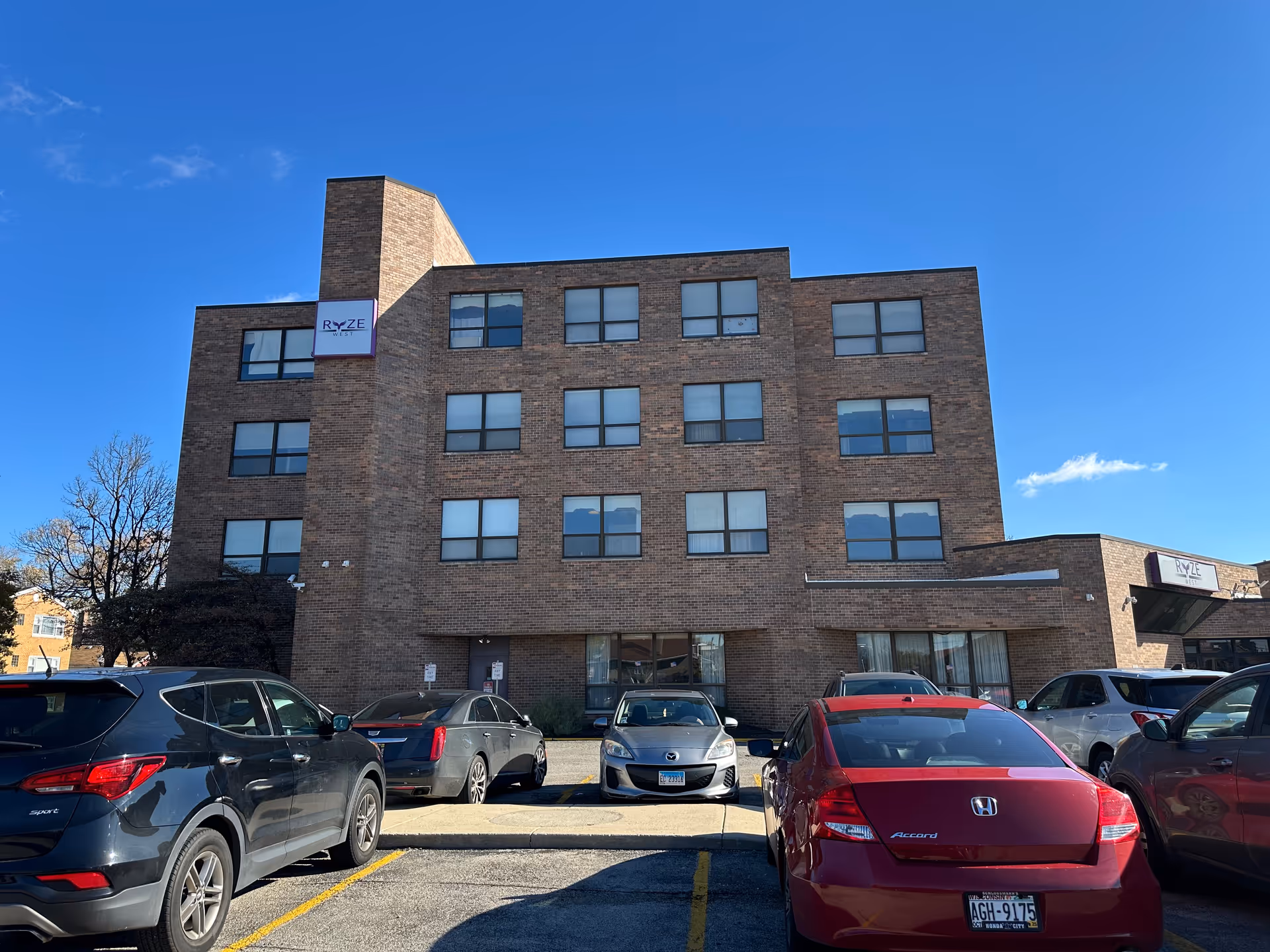 Brick four-story building labeled "RYZE West" with a parking lot and several cars in front under a clear blue sky.