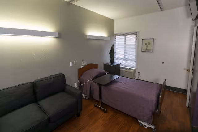 A small bedroom with a single hospital-style bed covered with a purple blanket, a dark gray couch, a small black bedside table with a plant, a window with blinds, and two wall-mounted lights above the bed.