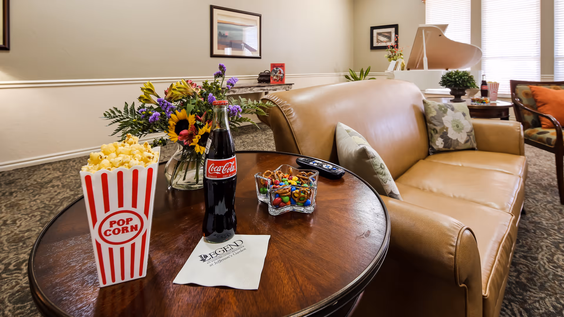 A cozy living room area with a tan leather sofa adorned with floral pillows. In front of the sofa is a round wooden table holding a striped popcorn container, a glass bottle of Coca-Cola, a bowl of mixed snacks, a TV remote, a vase with colorful flowers, and a napkin with the Legend At Jefferson's Garden logo. In the background, there is a white grand piano and a chair with an orange cushion near windows with blinds.