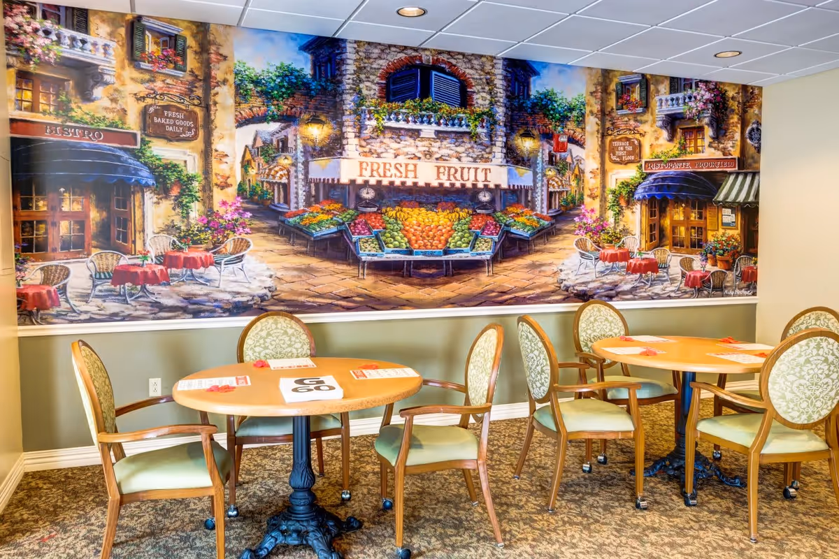 Interior dining area with round tables and green cushioned chairs on a patterned carpet. The wall features a large colorful mural depicting a European-style street market with fresh fruit stands, bistros, and outdoor seating.