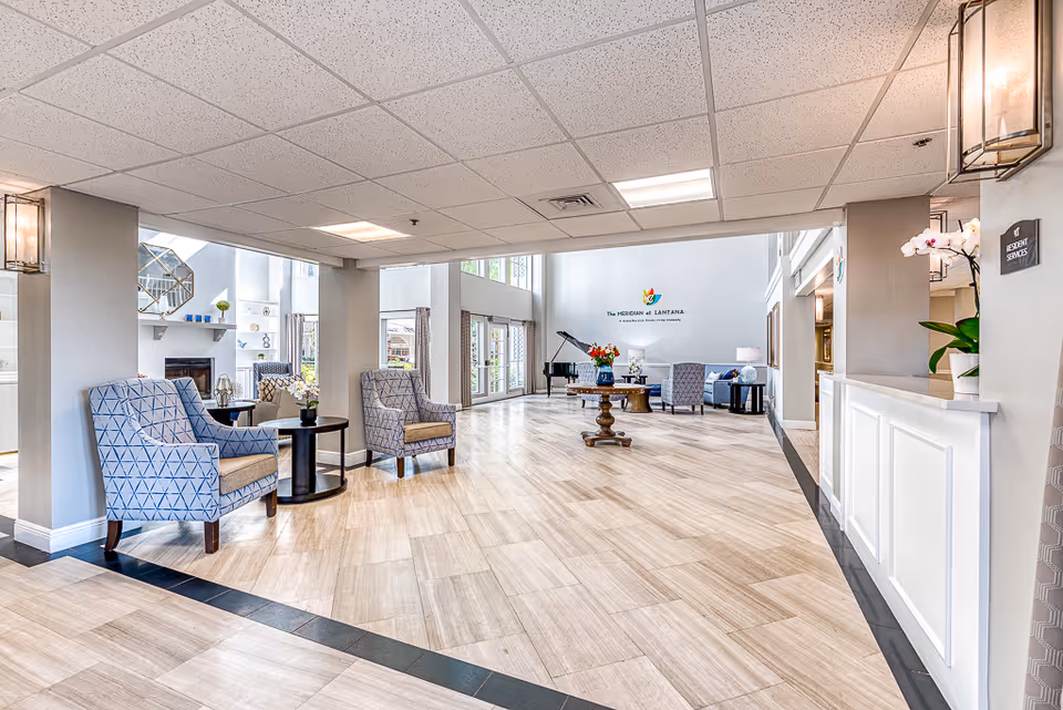 Bright and spacious senior living facility lobby with light wood flooring, patterned armchairs, small round tables with flowers, a grand piano, and a reception desk. The walls are painted light gray with large windows allowing natural light. A sign on the back wall reads 'The Meridian at Lantana'.