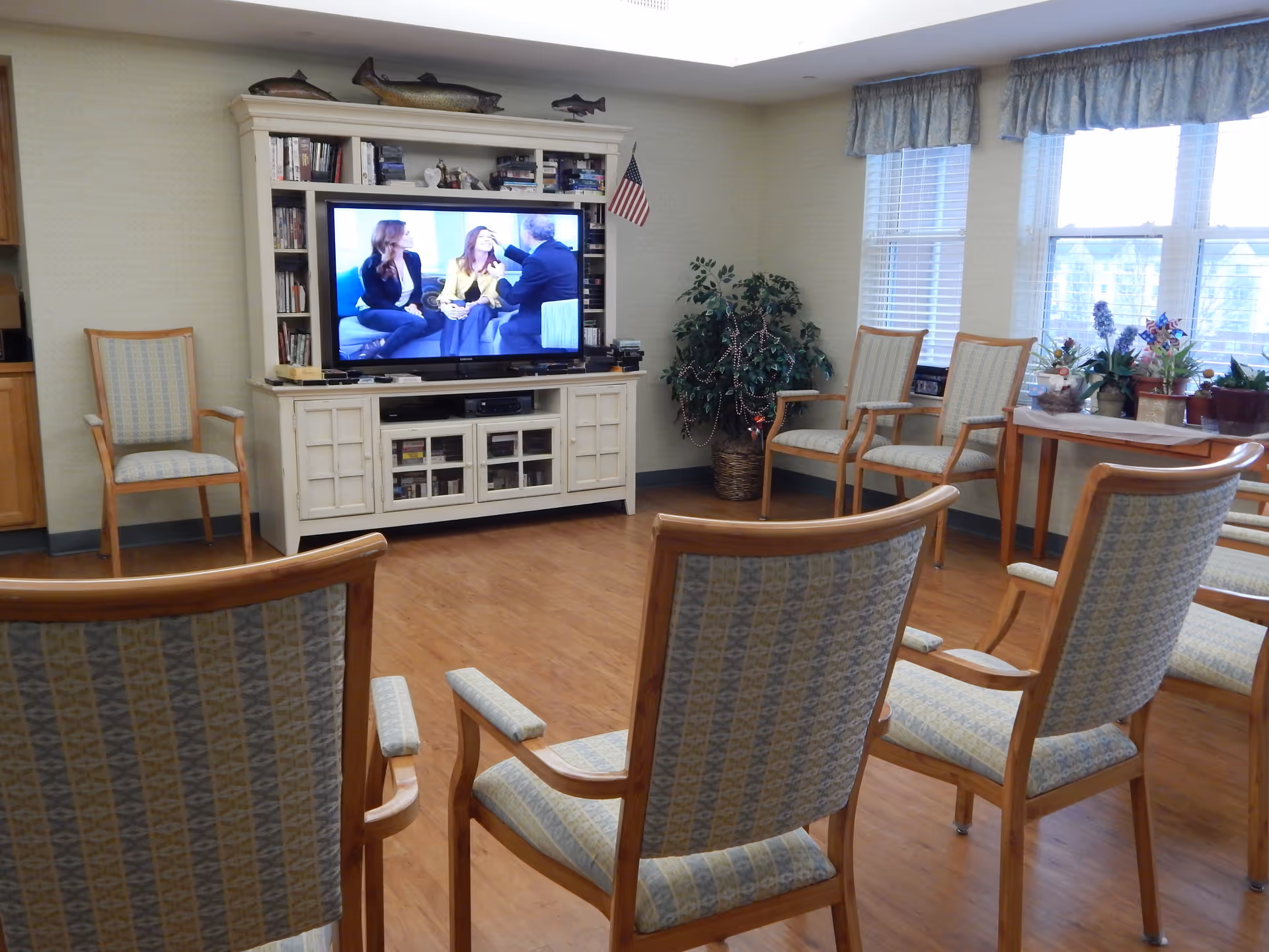 A cozy common room with several wooden chairs upholstered in patterned fabric arranged facing a large flat-screen TV on a white entertainment center. The TV is showing a talk show with three people. There is a small American flag on the entertainment center, a potted plant decorated with beads, and a table with various potted plants near windows with blue valance curtains.