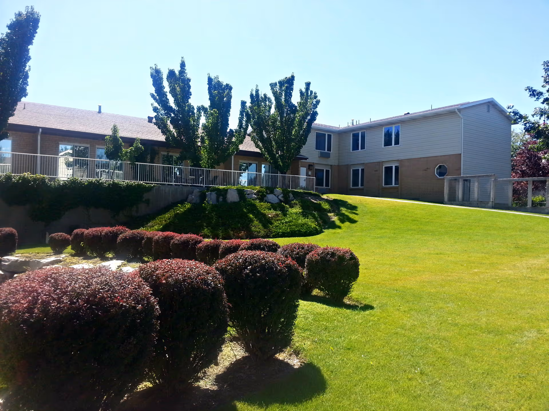 Exterior view of a two-story assisted living facility building with beige siding and brick accents. The building is surrounded by a well-maintained lawn, neatly trimmed bushes, and several tall trees. The sky is clear and blue.