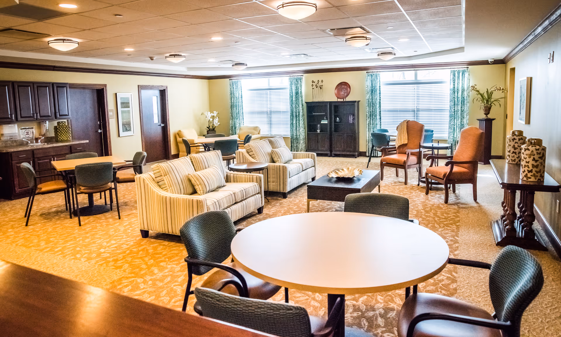 Bright communal lounge with sofas, armchairs, round tables and a small kitchenette in a senior living facility.