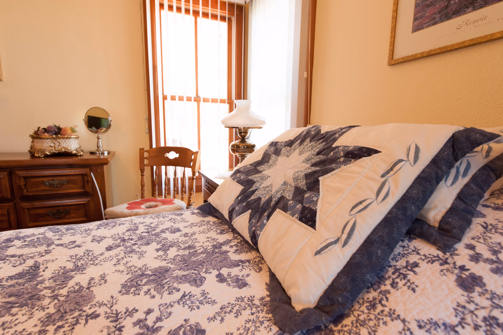 A cozy bedroom with a bed covered in a floral and star-patterned quilt and matching pillows. There is a wooden dresser with decorative items and a small round mirror, a wooden chair with a cushion, and a window with vertical blinds letting in natural light.