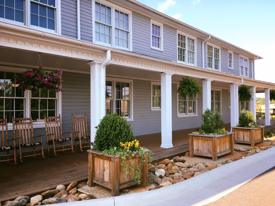 Exterior view of a senior living facility with a covered porch featuring wooden rocking chairs, hanging flower baskets, and large wooden planters with green plants and flowers. The building has gray siding, white columns, and multiple windows.