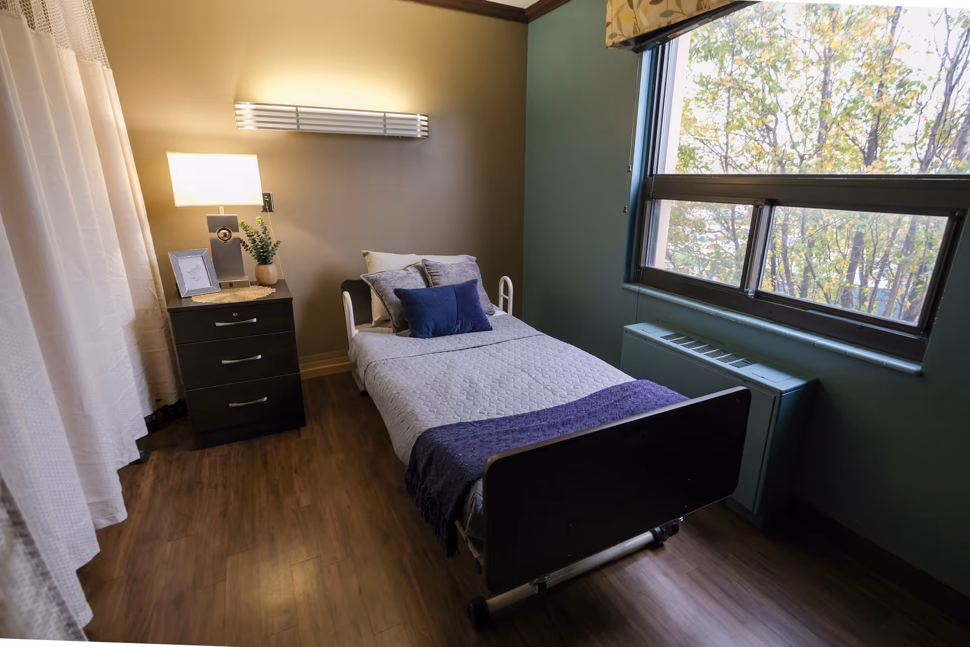 A small furnished bedroom with a single bed, nightstand and lamp beside a large window overlooking trees.