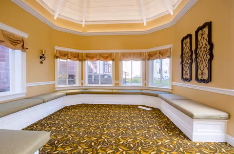 A bright, octagonal room with large windows on three sides, featuring built-in cushioned bench seating along the walls. The walls are painted a warm beige color with white trim, and the ceiling is vaulted with white beams. The floor is covered with a patterned carpet in shades of green, yellow, and brown. Two decorative wrought iron wall hangings are mounted on one wall.