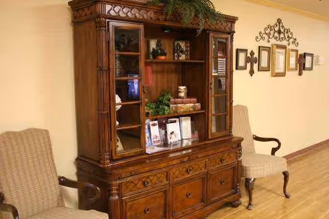 A tall antique wooden cabinet with glass doors flanked by two upholstered chairs in a hallway.