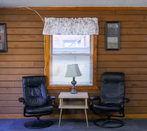 Two black recliner chairs sit beside a small table and lamp by a window.