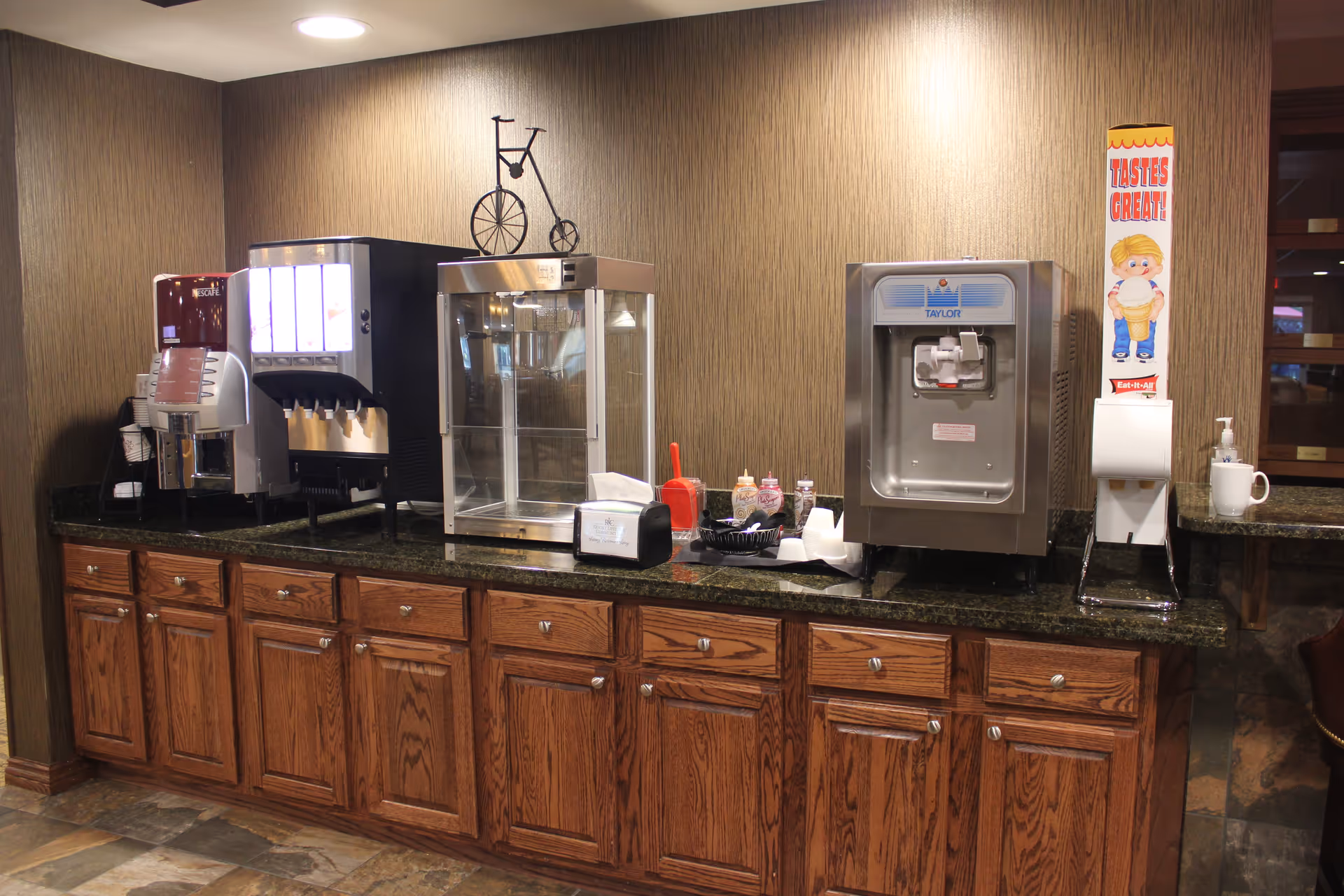 A countertop with wooden cabinets below, featuring a coffee machine, a soda dispenser, a popcorn machine, a soft serve ice cream machine, and various condiments and napkins. The wall behind is covered with textured wallpaper and there is a small sign that says 'TASTES GREAT!' with a cartoon character holding an ice cream cone.