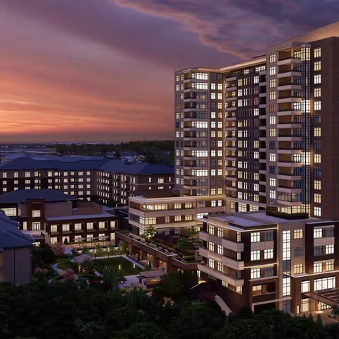 Exterior view of The Cardinal at North Hills senior living facility at dusk, showing multiple connected buildings with many lit windows and landscaped outdoor areas under a colorful sunset sky.